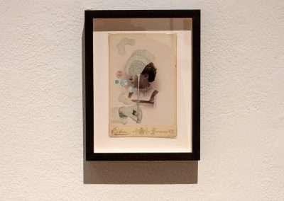 A framed mixed-media artwork featuring a vintage portrait with floral and abstract embellishments, mounted on a textured white wall.