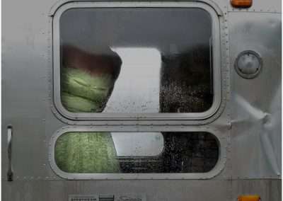A close-up of a rain-speckled Airstream trailer window, revealing a blurred view of a green cushion inside, set against a sleek silver exterior.