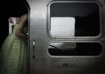 A woman in a pale green dress stands partially visible through the rounded door of a vintage silver trailer, facing away. The trailer's window reflects her figure and raindrops speckle its surface.