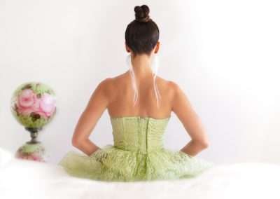 A woman in a green ballet costume with a tutu, seen from behind, sits against a white background. Her hair is styled in a bun, and a floral arrangement is visible to her side.