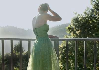 A woman in a green dress stands at a balcony railing, looking out over a forested landscape in the morning light. She has her hands in her hair, enjoying the tranquil view.