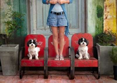 A woman in denim shorts and a blue cardigan stands holding a book, flanked by two small dogs seated on rustic red cinema chairs, against a colorful, aged wall.