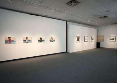 A modern art gallery with white walls featuring a variety of framed photographs hung at eye level. The spacious room has soft lighting and a grey carpeted floor.