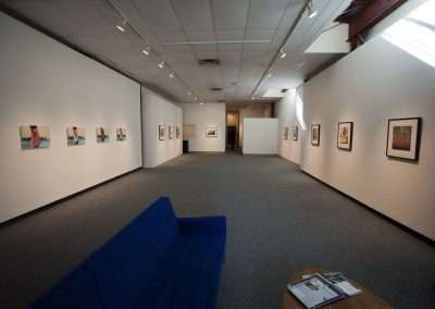 An art gallery interior with white walls displaying various framed artworks, illuminated by ceiling spotlights. A blue bench and a small table with reading material are in the foreground.