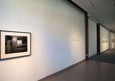 A bright gallery corridor displaying a framed photograph of a cactus on a TV, hung on a plain wall, with soft lighting and the shadowy outline of a person visible across other artworks.