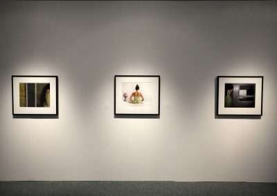 Three framed photographs displayed on a gallery wall, each illuminated by individual spotlights, with a central image featuring a person in white attire.