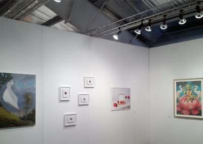 An art fair in Aspen with white walls displaying various paintings under bright ceiling lights. Some artworks feature realistic style while others are abstract.