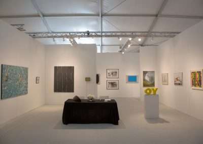 An art exhibition room with various paintings and framed works hung on white partition walls, featuring a central table with informational pamphlets and a white sculpture on a pedestal.