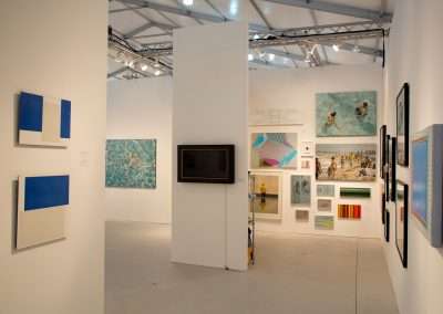 Art gallery interior showing a variety of modern art pieces, including abstract paintings and a photograph, displayed on white walls under bright lighting.