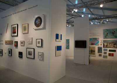 An art exhibition space with various framed artworks displayed on white walls under bright lighting, including paintings, photographs, and mixed-media pieces.