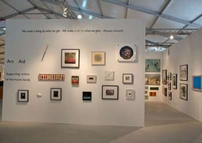 An art exhibit featuring various styles of artwork, including paintings and photographs, displayed on white walls under gallery lighting. A quote by Winston Churchill is at the top: "We make a living by what we get. We make a life by what we give.