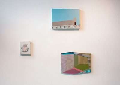 Three abstract paintings on a white gallery wall, comprising a small DIY painting of a coffee cup, a larger canvas of a minimalist building, and a vibrant, geometric piece.