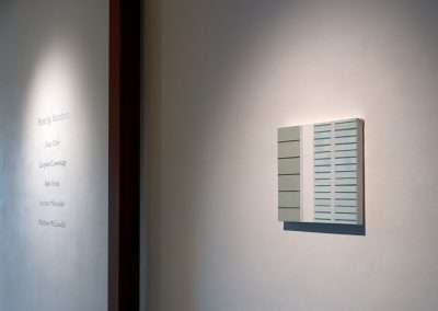White wall in an art gallery with a minimalist piece resembling a small window shutter, and a label listing various artist names next to it. A DIY painting kit lies nearby, hinting at interactive art. Soft lighting casts a gentle shadow on the wall.
