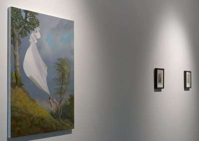A painting displayed in a gallery showing a surreal scene of a white sheet caught in a tree branch, with a seemingly headless figure beneath it; two smaller paint by numbers pieces hang on the adjacent wall.