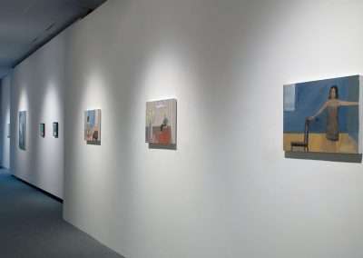 A modern art gallery hallway featuring a series of DIY paintings on the white walls, evenly spaced and illuminated by ceiling lights, with a blue carpet extending along the corridor.