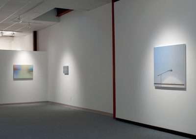 Gallery interior with white walls displaying three DIY paintings under soft lighting, one centered on a standalone panel and two on adjacent walls.