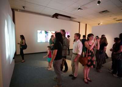 A diverse group of people, including adults and a child, view projected artistic images in a softly lit gallery space. Some attendees are capturing the moment with their cameras.