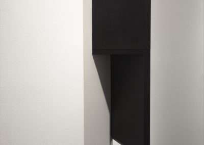 A modern geometric wall sculpture featuring a combination of angular shapes in black and cream colors, set against a white gallery wall.