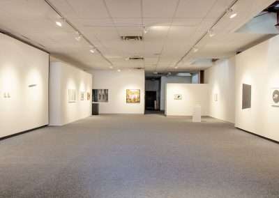 Interior of an art gallery showing an array of framed paintings on white walls, gray carpeted floors, and recessed lighting, creating a spacious and modern exhibit space.