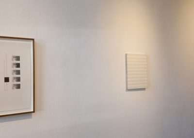 Two framed artworks on a gallery wall, one with a series of black and white photos and the other a white textured piece, under the inscription "20 in 2011.
