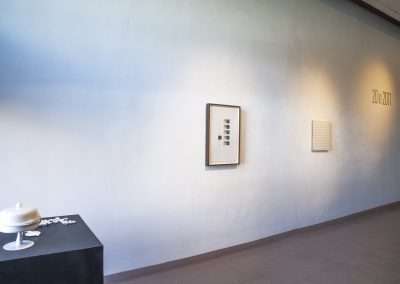 A minimalist art gallery installation with a white urn on a black pedestal, scattered white petals, and two framed artworks on a light gray wall lit by natural light.