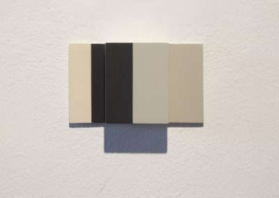 A minimalist artwork displayed on a white wall, featuring a series of adjacent panels in black, dark gray, light gray, and beige. The shadow beneath the artwork creates a subtle depth effect.