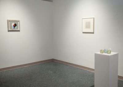 Art gallery interior with two white walls displaying framed artworks and a pedestal with glass sculptures. Simple and minimalist aesthetic.