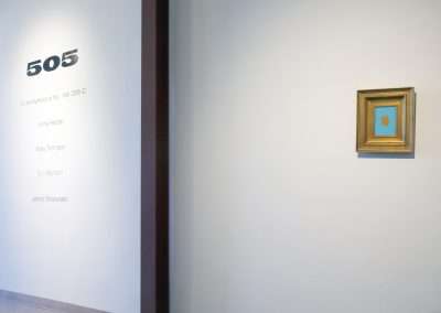 A minimalist art gallery wall featuring a small framed painting of a blue circle centered on an orange square, next to a doorway with the number 505 and a list of artist names.