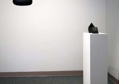 A minimalist art gallery installation featuring a small black sculpture on a white pedestal, with a dark, elliptical object suspended on the white wall above it.