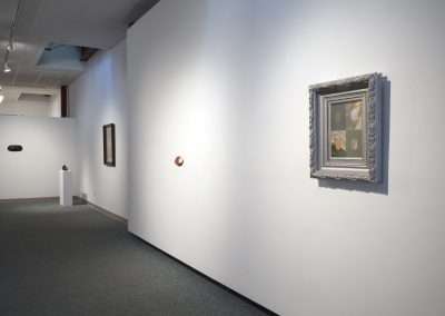 An art gallery room with white walls displaying various framed artworks, including a prominently featured ornate silver frame with a modern painting.