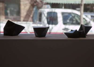 Three black origami boats lined up on a white ledge, with a blurred street scene in the background featuring a white van.