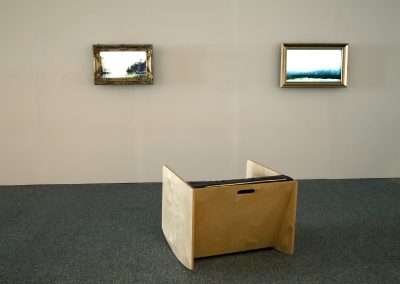 A minimalist art gallery room with two landscape paintings hung on a light grey wall and an abstract, low, cubic chair-like sculpture positioned centrally on a grey carpeted floor.