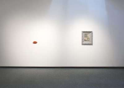 A minimalist art gallery with a plain white wall featuring a small framed picture on the right and an egg-shaped object floating near the center-left.