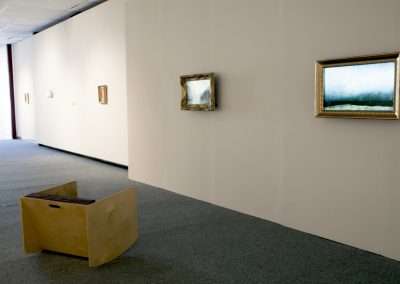 A modern art gallery with beige walls featuring several landscape paintings, and a square wooden bench placed at the center on a gray carpeted floor.