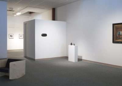 An art gallery room with white walls displaying various framed artworks, a sculpture on a pedestal, and a bench on the left for viewing.