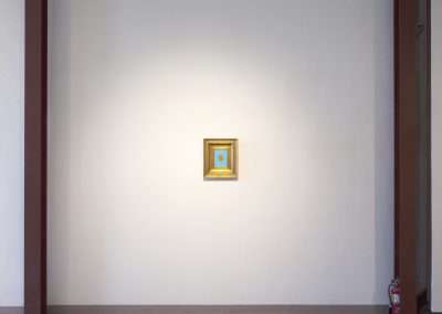 A small, framed golden artwork centered on a large, plain white wall, flanked by brown edges, with a fire extinguisher at the bottom right corner.