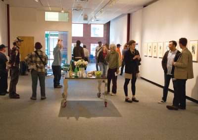 People mingling at an art gallery opening, viewing framed artworks on walls, and conversing while standing near a refreshment table.