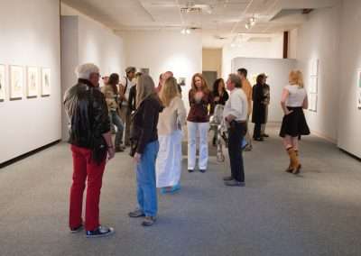 People viewing and discussing various artworks at a well-lit art gallery exhibition, featuring walls adorned with colorful paintings and photographs.