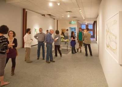 People conversing and viewing artwork at an art gallery opening reception. The room is well-lit with white walls and artwork hung for display.