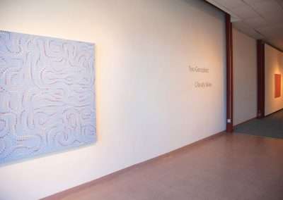 Art gallery interior with a large abstract painting titled "Cloudy Skies" by Teo Gonzalez displayed on a beige wall, featuring intricate dot patterns.