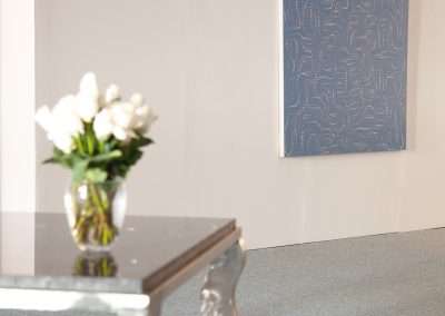 A modern art gallery scene with a clear acrylic table holding a vase of white flowers, set against a textured blue painting with an intricate white line pattern on the wall.