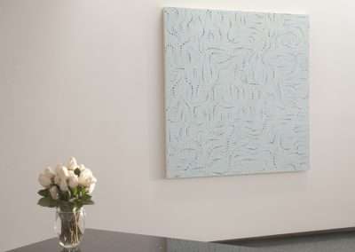 A modern art painting featuring a complex blue and white pattern hangs on a white gallery wall next to a glass vase with white flowers on a reflective metal table.