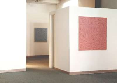 Art gallery interior showing abstract textured paintings hanging on a white wall, with sunlight casting soft shadows on the floor.