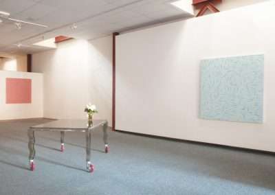 An art gallery with minimalistic decor featuring two large abstract paintings on white walls and a small metal table with wheels holding a white vase.