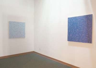 Two abstract paintings with swirling patterns, one in shades of blue and the other in blue and white, displayed on the walls of a bright, empty gallery room.