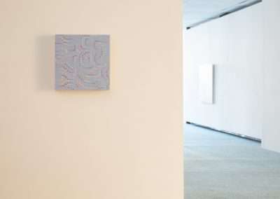 A modern art gallery with plain walls and carpeted floor, featuring a small, square abstract painting with blue tones hanging on the left wall.
