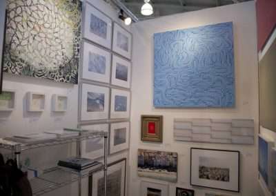An art exhibition booth displaying various artworks, including a prominent blue abstract piece, surrounded by other mixed media works and paintings on white walls.