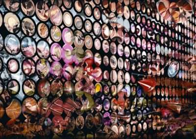 A colorful wall installation composed of numerous circular discs, each displaying different intricate patterns, illuminated dramatically in a dark setting.