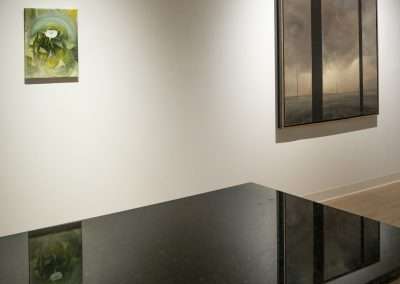A modern art gallery interior featuring two paintings on a beige wall, one abstract in greens and yellows, and one realistic showing a stormy sky. A reflective black surface in the foreground adds depth.