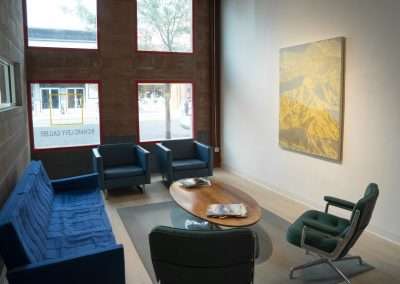 A modern office lobby with contemporary furniture, including blue and green chairs, and a glass door leading outside. A large painting of a mountain hangs on the wall.